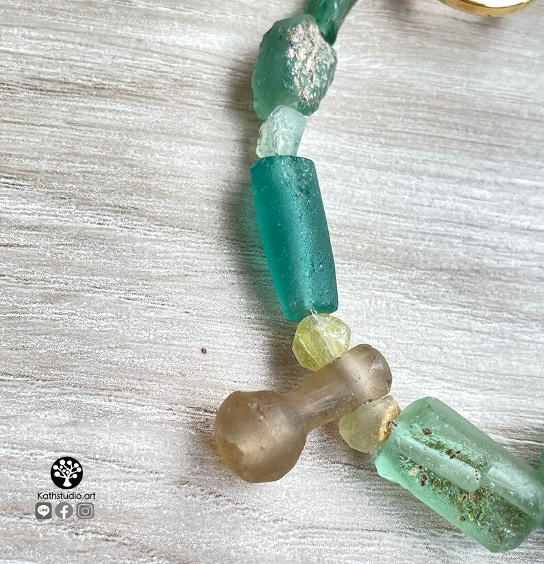 Puede incluir: Primer plano de un collar con cuentas de vidrio marino de varias formas y tama&ntilde;os, en tonos verdes, azules y amarillos. Las cuentas est&aacute;n ensartadas, creando un dise&ntilde;o &uacute;nico y org&aacute;nico. El collar est&aacute; sobre una superficie de madera clara.