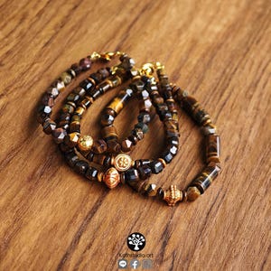 May include: Three bracelets crafted from faceted tiger eye beads in shades of brown and gold. Each bracelet features a central gold-toned metal charm with intricate detailing and a gold-toned lobster claw clasp closure for secure fastening.
