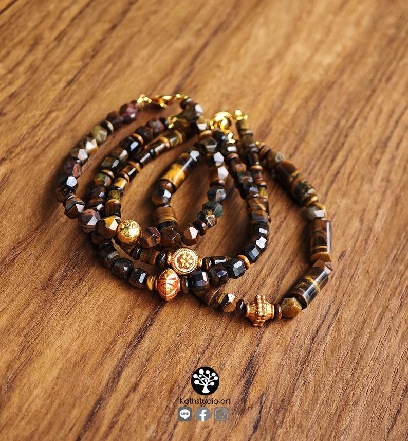 May include: Three bracelets crafted from faceted tiger eye beads in shades of brown and gold. Each bracelet features a central gold-toned metal charm with intricate detailing and a gold-toned lobster claw clasp closure for secure fastening.