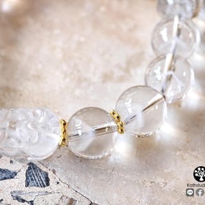 May include: A bracelet featuring clear, spherical quartz beads and a carved, translucent creature bead. The beads are separated by small, gold-toned metal spacers. The logo Kathstudio.art is visible in the bottom right corner of the image.