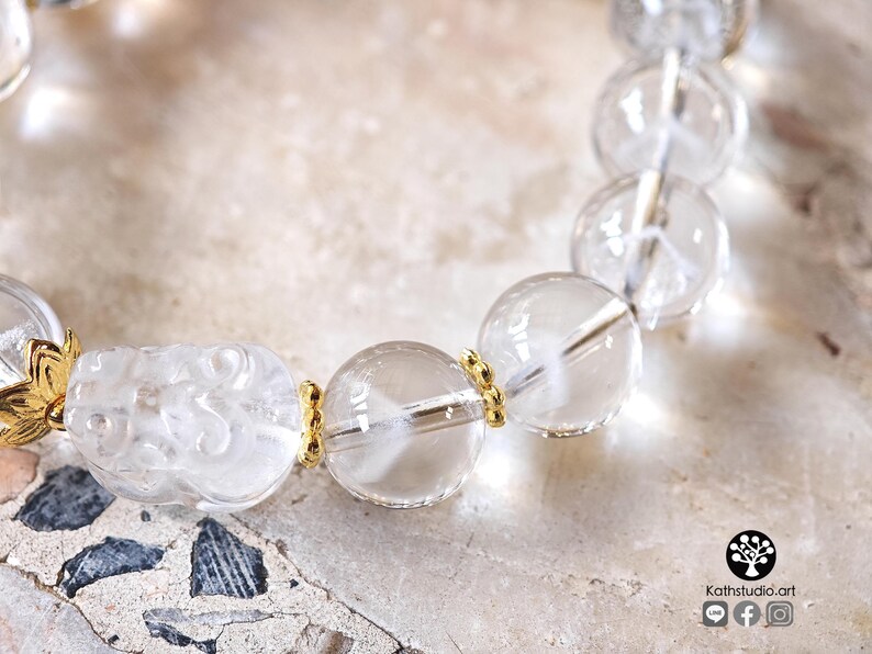 May include: A bracelet featuring clear, spherical quartz beads and a carved, translucent creature bead. The beads are separated by small, gold-toned metal spacers. The logo Kathstudio.art is visible in the bottom right corner of the image.