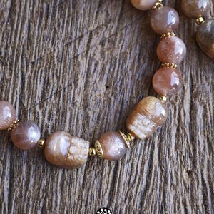 May include: A beaded bracelet featuring round, polished sunstone beads in shades of peach and tan, interspersed with small gold-toned metal spacers and two carved sunstone skull beads. The logo Kathstudio.art is printed at the bottom.