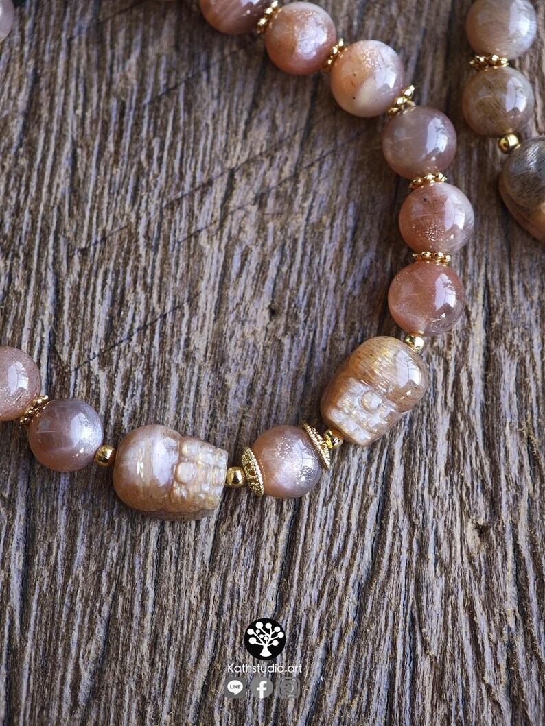 May include: A beaded bracelet featuring round, polished sunstone beads in shades of peach and tan, interspersed with small gold-toned metal spacers and two carved sunstone skull beads. The logo Kathstudio.art is printed at the bottom.