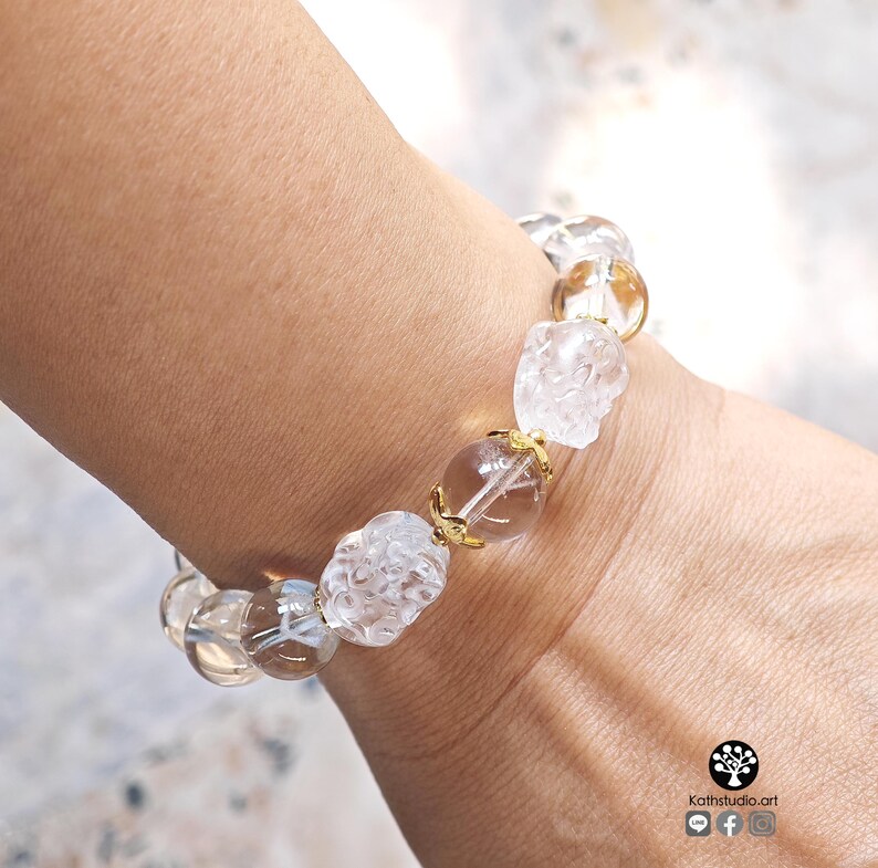 May include: A beaded bracelet featuring clear, spherical quartz beads and carved, textured clear quartz beads. The beads are accented with small, gold-toned metal spacers. The bracelet is worn on a wrist, and a small logo with the text Kathstudio.art is visible.