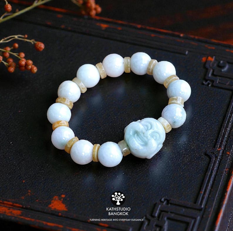Op de afbeelding: Een kralenarmband met gladde, bleekwitte jadebollen afgewisseld met kleine, doorschijnende lichtbruine tussenkralen. Een grotere, uitgesneden lichtgroene jade bedel in de vorm van een lachend gezicht is in het ontwerp verwerkt.
