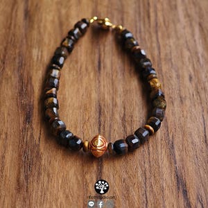 May include: A beaded bracelet featuring faceted dark brown and golden tiger eye stones arranged in a circular pattern. A single, central, polished gold-toned metal bead with an embossed geometric eye design serves as the focal point of the piece.