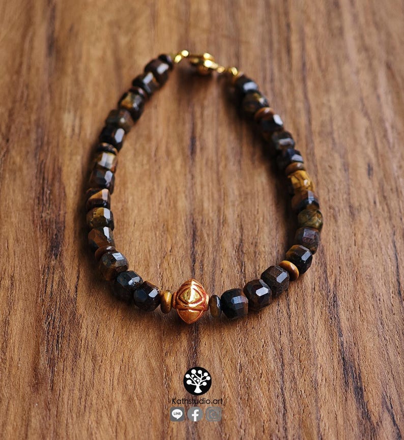 May include: A beaded bracelet featuring faceted dark brown and golden tiger eye stones arranged in a circular pattern. A single, central, polished gold-toned metal bead with an embossed geometric eye design serves as the focal point of the piece.