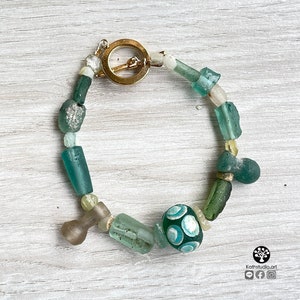 Puede incluir: Una pulsera hecha a mano con una variedad de cuentas de vidrio en tonos verdes, azules y transparentes. La pulsera incluye una cuenta grande y redonda de color verde oscuro con un patr&oacute;n azul claro. El cierre es dorado.