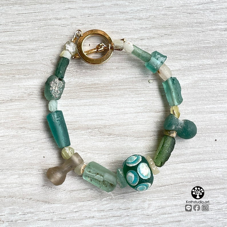 Puede incluir: Una pulsera hecha a mano con una variedad de cuentas de vidrio en tonos verdes, azules y transparentes. La pulsera incluye una cuenta grande y redonda de color verde oscuro con un patr&oacute;n azul claro. El cierre es dorado.