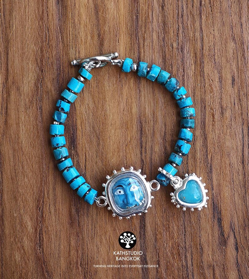 Puede incluir: Una pulsera con cuentas cil&iacute;ndricas de turquesa ensartadas en una cadena de plata con cierre de barra. La pieza incluye un dije central enmarcado en plata que representa un rostro azul y un colgante de turquesa en forma de coraz&oacute;n.