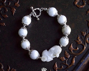 May include: A bracelet featuring round white gemstone beads accented with ornate silver-toned metal caps and a central carved white stone figure. The piece includes a silver-toned toggle clasp and is marked with the text KATHSTUDIO BANGKOK.