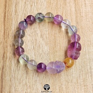 May include: A stretch bracelet composed of round, translucent fluorite beads in shades of purple, green, and yellow, interspersed with two carved skull-shaped beads. The beads are strung on a thin, dark purple elastic cord, creating a circular design.