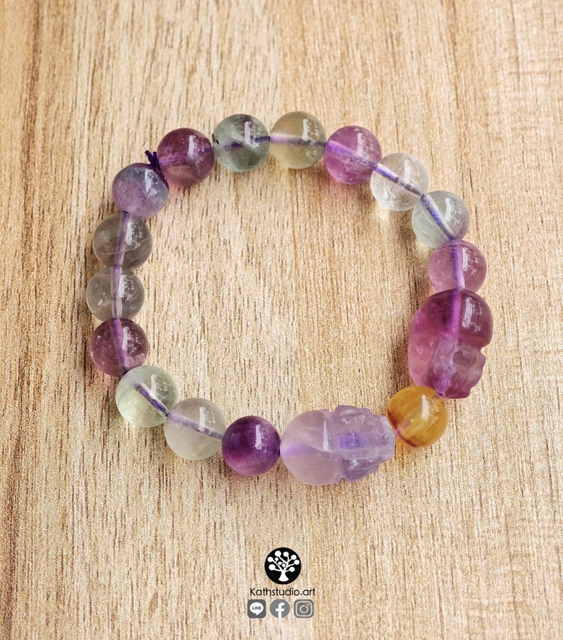 May include: A stretch bracelet composed of round, translucent fluorite beads in shades of purple, green, and yellow, interspersed with two carved skull-shaped beads. The beads are strung on a thin, dark purple elastic cord, creating a circular design.