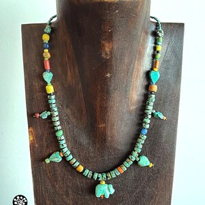 Op de afbeelding: Een turquoise kralenketting met een verscheidenheid aan kleurrijke kralen en bedels, waaronder hartvormige en olifantenhangers. De ketting wordt tentoongesteld op een houten standaard. De ketting is gemaakt van turquoise, gele, oranje en zwarte kralen.