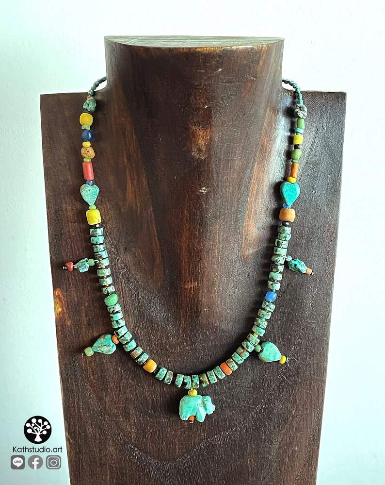 Op de afbeelding: Een turquoise kralenketting met een verscheidenheid aan kleurrijke kralen en bedels, waaronder hartvormige en olifantenhangers. De ketting wordt tentoongesteld op een houten standaard. De ketting is gemaakt van turquoise, gele, oranje en zwarte kralen.