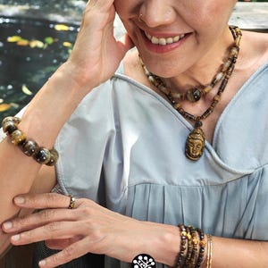 Puede incluir: Un collar de cuentas de ojo de tigre con un colgante de Buda tallado, una pulsera de cuentas a juego y un conjunto de pulseras finas. El juego de joyas incluye piedras marrones y doradas con vetas naturales, usado con una blusa gris claro.