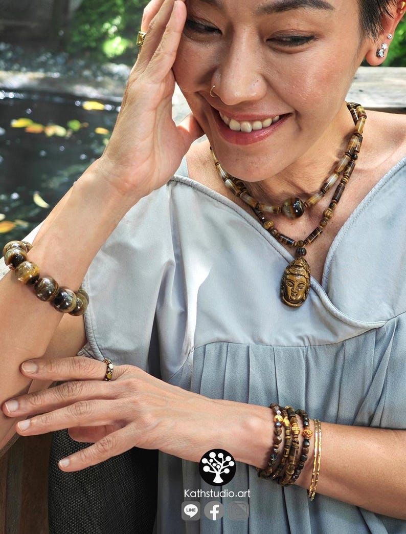 Puede incluir: Un collar de cuentas de ojo de tigre con un colgante de Buda tallado, una pulsera de cuentas a juego y un conjunto de pulseras finas. El juego de joyas incluye piedras marrones y doradas con vetas naturales, usado con una blusa gris claro.