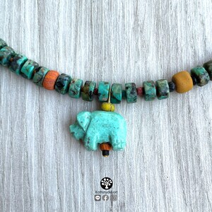 Op de afbeelding: Een turquoise kralenketting met een olifantenhanger. De ketting heeft turquoise, oranje en bruine kralen. De olifantenhanger is lichtblauw van kleur. De ketting wordt gepresenteerd op een licht houten oppervlak.