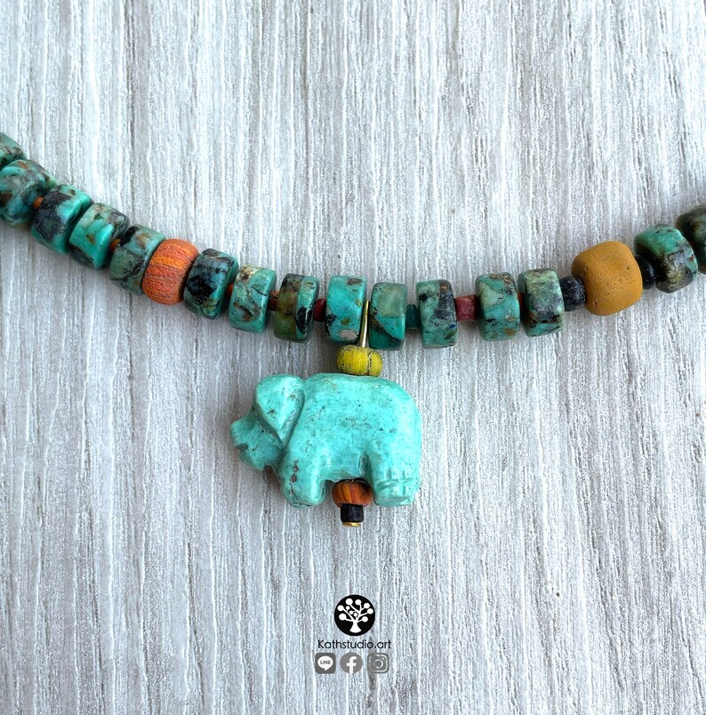 Op de afbeelding: Een turquoise kralenketting met een olifantenhanger. De ketting heeft turquoise, oranje en bruine kralen. De olifantenhanger is lichtblauw van kleur. De ketting wordt gepresenteerd op een licht houten oppervlak.