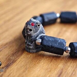 May include: A bracelet featuring a carved dark gray stone animal bead with bright red inlaid eyes, interspersed with rectangular matte black beads and small metallic silver-toned faceted spacer beads arranged in a circular pattern.