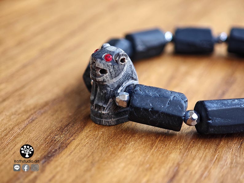 May include: A bracelet featuring a carved dark gray stone animal bead with bright red inlaid eyes, interspersed with rectangular matte black beads and small metallic silver-toned faceted spacer beads arranged in a circular pattern.