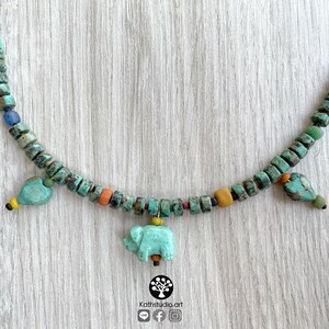 Op de afbeelding: Een turquoise kralenketting met een olifantenhanger en hartvormige bedels. De ketting heeft een verscheidenheid aan kleurrijke kralen, waaronder turquoise, oranje, geel en bruin. De ketting wordt gepresenteerd op een licht houten oppervlak. Het logo van de winkel is rechtsonder zichtbaar.