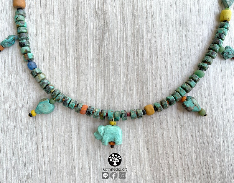Op de afbeelding: Een turquoise kralenketting met een olifantenhanger en hartvormige bedels. De ketting heeft een verscheidenheid aan kleurrijke kralen, waaronder turquoise, oranje, geel en bruin. De ketting wordt gepresenteerd op een licht houten oppervlak. Het logo van de winkel is rechtsonder zichtbaar.