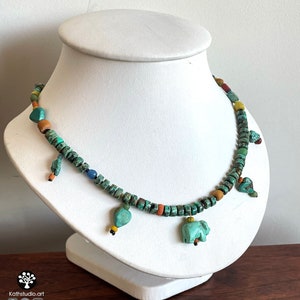 Op de afbeelding: Een turquoise kralenketting met kleine, vierkante kralen en verschillende bungelende bedels. De bedels zijn onder meer hartvormen en een kleine olifant. De ketting wordt op een witte buste getoond.