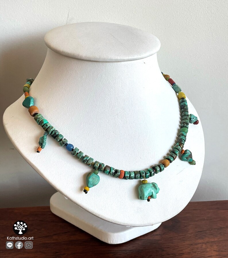 Op de afbeelding: Een turquoise kralenketting met kleine, vierkante kralen en verschillende bungelende bedels. De bedels zijn onder meer hartvormen en een kleine olifant. De ketting wordt op een witte buste getoond.