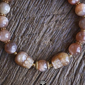 May include: A beaded bracelet featuring round, translucent peach-toned sunstone beads and two carved skull-shaped sunstone beads. The beads are separated by small, metallic gold-colored spacers. The text Kathstudio.art is printed at the bottom.
