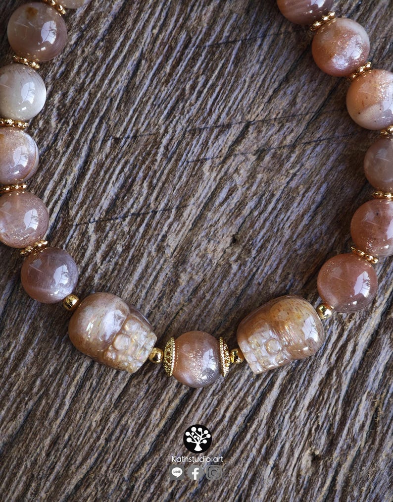 May include: A beaded bracelet featuring round, translucent peach-toned sunstone beads and two carved skull-shaped sunstone beads. The beads are separated by small, metallic gold-colored spacers. The text Kathstudio.art is printed at the bottom.
