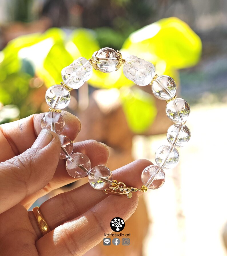 May include: A bracelet featuring clear, spherical crystal beads interspersed with gold-toned metal spacers and two carved, translucent crystal figures. The design includes a gold-toned metal clasp closure. The image includes the text Kathstudio.art.