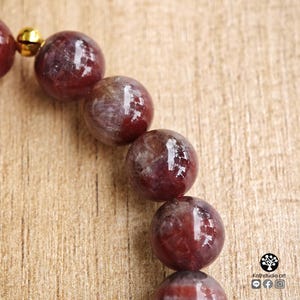 Peut inclure: Un bracelet compos&eacute; de perles sph&eacute;riques en pierre gemme rouge fonc&eacute; polies, pr&eacute;sentant des marbrures et des inclusions internes visibles. Une petite clochette en m&eacute;tal dor&eacute; brillant est fix&eacute;e au bracelet, pr&egrave;s du haut de la rang&eacute;e de perles.