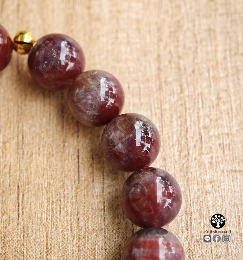 Peut inclure: Un bracelet compos&eacute; de perles sph&eacute;riques en pierre gemme rouge fonc&eacute; polies, pr&eacute;sentant des marbrures et des inclusions internes visibles. Une petite clochette en m&eacute;tal dor&eacute; brillant est fix&eacute;e au bracelet, pr&egrave;s du haut de la rang&eacute;e de perles.