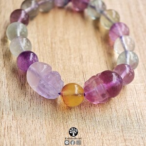 May include: A beaded bracelet featuring translucent fluorite spheres in shades of purple, green, and clear, interspersed with two carved skull-shaped beads and a single round yellow bead. The text Kathstudio.art is printed at the bottom.