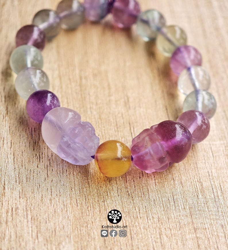 May include: A beaded bracelet featuring translucent fluorite spheres in shades of purple, green, and clear, interspersed with two carved skull-shaped beads and a single round yellow bead. The text Kathstudio.art is printed at the bottom.