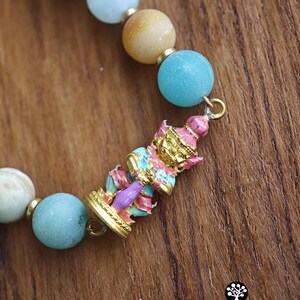 May include: A beaded bracelet featuring round light blue and tan stones separated by small gold-toned spacers. A central gold-toned charm depicts a standing figure painted in pink, light blue, and gold. The text Kathstudio.art is visible.