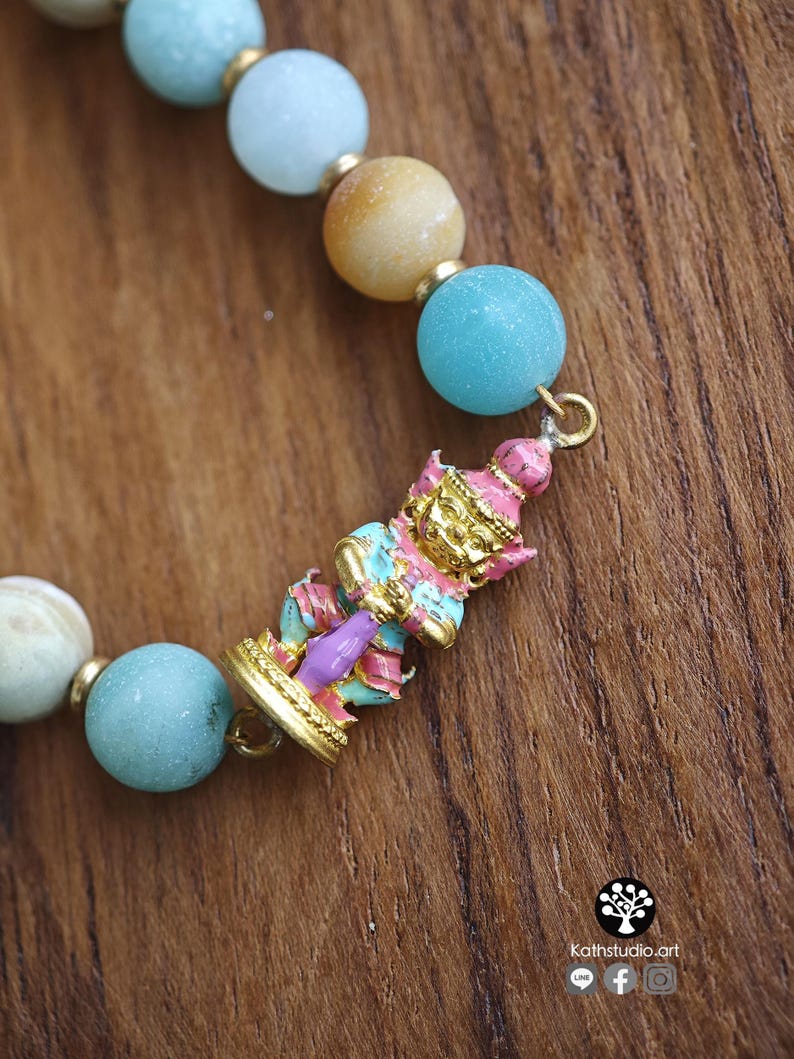 May include: A beaded bracelet featuring round light blue and tan stones separated by small gold-toned spacers. A central gold-toned charm depicts a standing figure painted in pink, light blue, and gold. The text Kathstudio.art is visible.