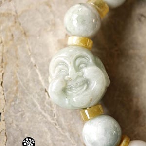 Op de afbeelding: Een kralenarmband met gepolijste, lichtgroene jade bollen en kleinere, cilindrische gele jade tussenstukjes. Een centrale, uitgesneden lichtgroene jade kraal toont een lachend Boeddha-gezicht. Het logo KATHSTUDIO BANGKOK NG HERITAGE INTO EVERYDAY ELEGANCE is zichtbaar.