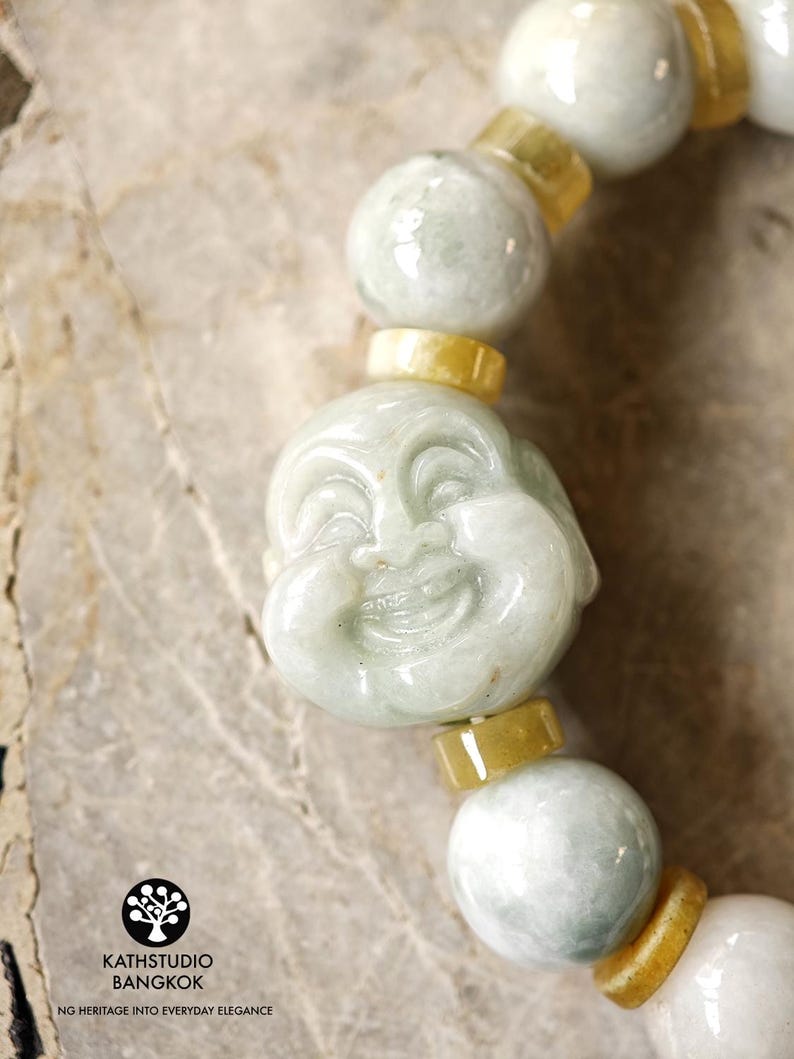 Op de afbeelding: Een kralenarmband met gepolijste, lichtgroene jade bollen en kleinere, cilindrische gele jade tussenstukjes. Een centrale, uitgesneden lichtgroene jade kraal toont een lachend Boeddha-gezicht. Het logo KATHSTUDIO BANGKOK NG HERITAGE INTO EVERYDAY ELEGANCE is zichtbaar.
