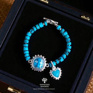Puede incluir: Una pulsera con cuentas cil&iacute;ndricas de turquesa, un cierre de palanca plateado, un dije rectangular con una ilustraci&oacute;n de rostro azul y un dije de coraz&oacute;n de turquesa. El texto KATHSTUDIO BANGKOK TURNING HERITAGE INTO EVERYDAY ELEGANCE es visible.