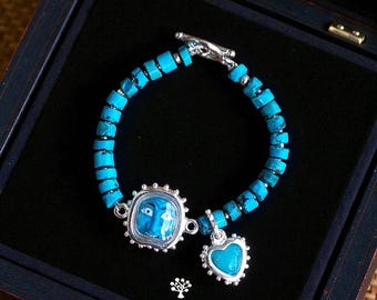 May include: A bracelet featuring cylindrical turquoise beads, a silver-toned toggle clasp, a rectangular charm with a blue face illustration, and a heart-shaped turquoise charm. The text KATHSTUDIO BANGKOK TURNING HERITAGE INTO EVERYDAY ELEGANCE is visible.