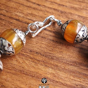 Puede incluir: Una pulsera con cuentas redondas y transl&uacute;cidas de color &aacute;mbar, separadas por tapas de metal plateado oxidado y ornamentado. La pieza se asegura con un mecanismo de cierre de gancho plateado. El texto Kathstudio.art es visible en la parte inferior.