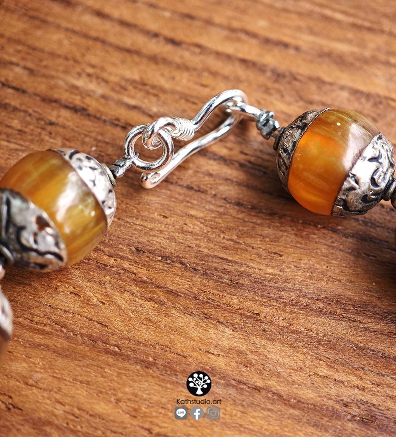 Puede incluir: Una pulsera con cuentas redondas y transl&uacute;cidas de color &aacute;mbar, separadas por tapas de metal plateado oxidado y ornamentado. La pieza se asegura con un mecanismo de cierre de gancho plateado. El texto Kathstudio.art es visible en la parte inferior.