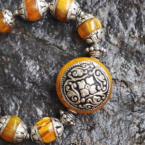 Puede incluir: Una pulsera con cuentas redondas y transl&uacute;cidas de color &aacute;mbar que se alternan con espaciadores de metal plateado ornamentados. Una cuenta circular central grande est&aacute; incrustada con una placa de metal plateado con motivos florales.