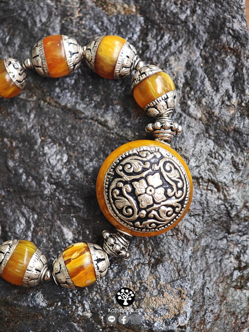 Puede incluir: Una pulsera con cuentas redondas y transl&uacute;cidas de color &aacute;mbar que se alternan con espaciadores de metal plateado ornamentados. Una cuenta circular central grande est&aacute; incrustada con una placa de metal plateado con motivos florales.