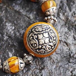 Puede incluir: Una pulsera con cuentas redondas de color &aacute;mbar veteado que se alternan con espaciadores de metal plateado ornamentado. La cuenta central es una pieza grande, plana y circular de color &aacute;mbar con un medall&oacute;n floral plateado en relieve.