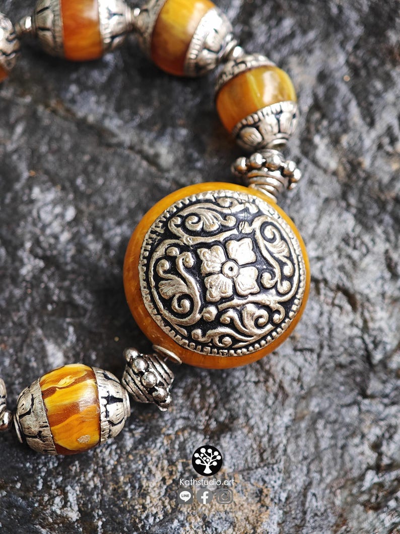 Puede incluir: Una pulsera con cuentas redondas de color &aacute;mbar veteado que se alternan con espaciadores de metal plateado ornamentado. La cuenta central es una pieza grande, plana y circular de color &aacute;mbar con un medall&oacute;n floral plateado en relieve.