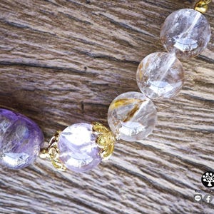 Peut inclure: Un bracelet de perles compos&eacute; de perles en am&eacute;thyste violette translucide en forme de cr&acirc;ne et de sph&egrave;res en quartz clair avec des inclusions dor&eacute;es. Les perles sont s&eacute;par&eacute;es par des capuchons en m&eacute;tal dor&eacute;. Logo avec Kathstudio.art visible.