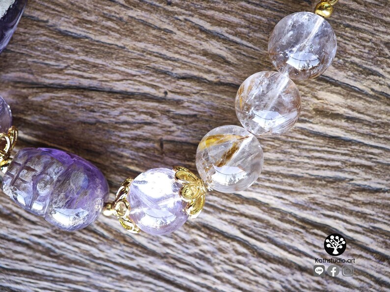 Peut inclure: Un bracelet de perles compos&eacute; de perles en am&eacute;thyste violette translucide en forme de cr&acirc;ne et de sph&egrave;res en quartz clair avec des inclusions dor&eacute;es. Les perles sont s&eacute;par&eacute;es par des capuchons en m&eacute;tal dor&eacute;. Logo avec Kathstudio.art visible.